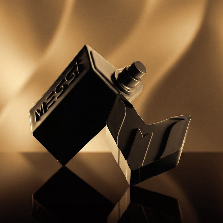 A black rectangular perfume bottle with a matching black cap, shown at an angle on a reflective dark surface against a warm golden-brown background. The bottle has the brand name 'Messi' embossed on its side.